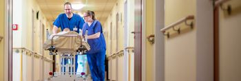 Zwei Pflegekräfte schieben einen Patienten auf einem mobilen Pflegebett den Gang entlang