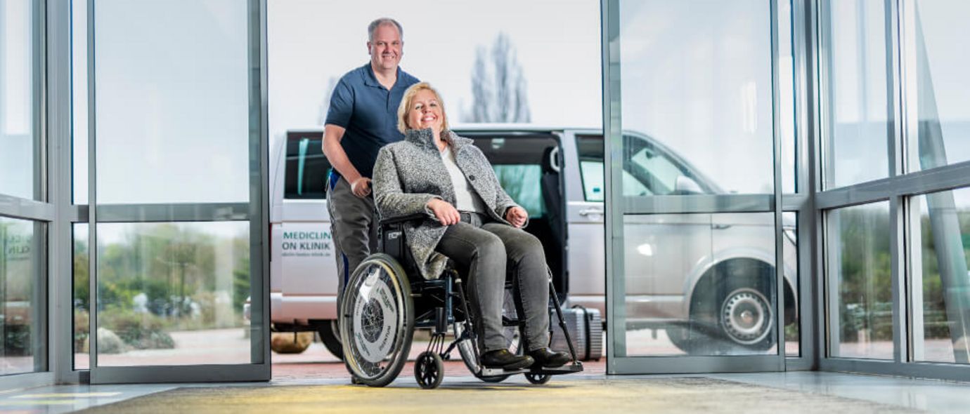 Mitarbeiter des Bringdienstes schiebt einen Patienten im Rollstuhl durch die Eingangstür der Klinik
