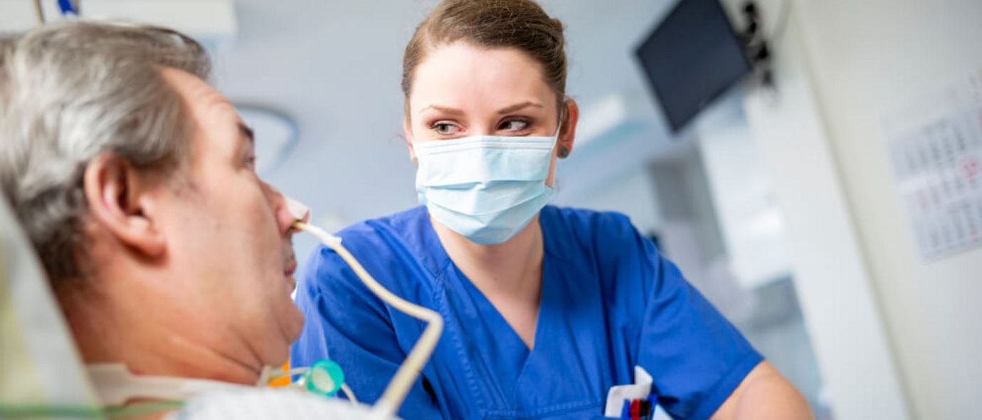Pflegekraft mit Maske spricht mit Neurologie-Patienten am Krankenbett